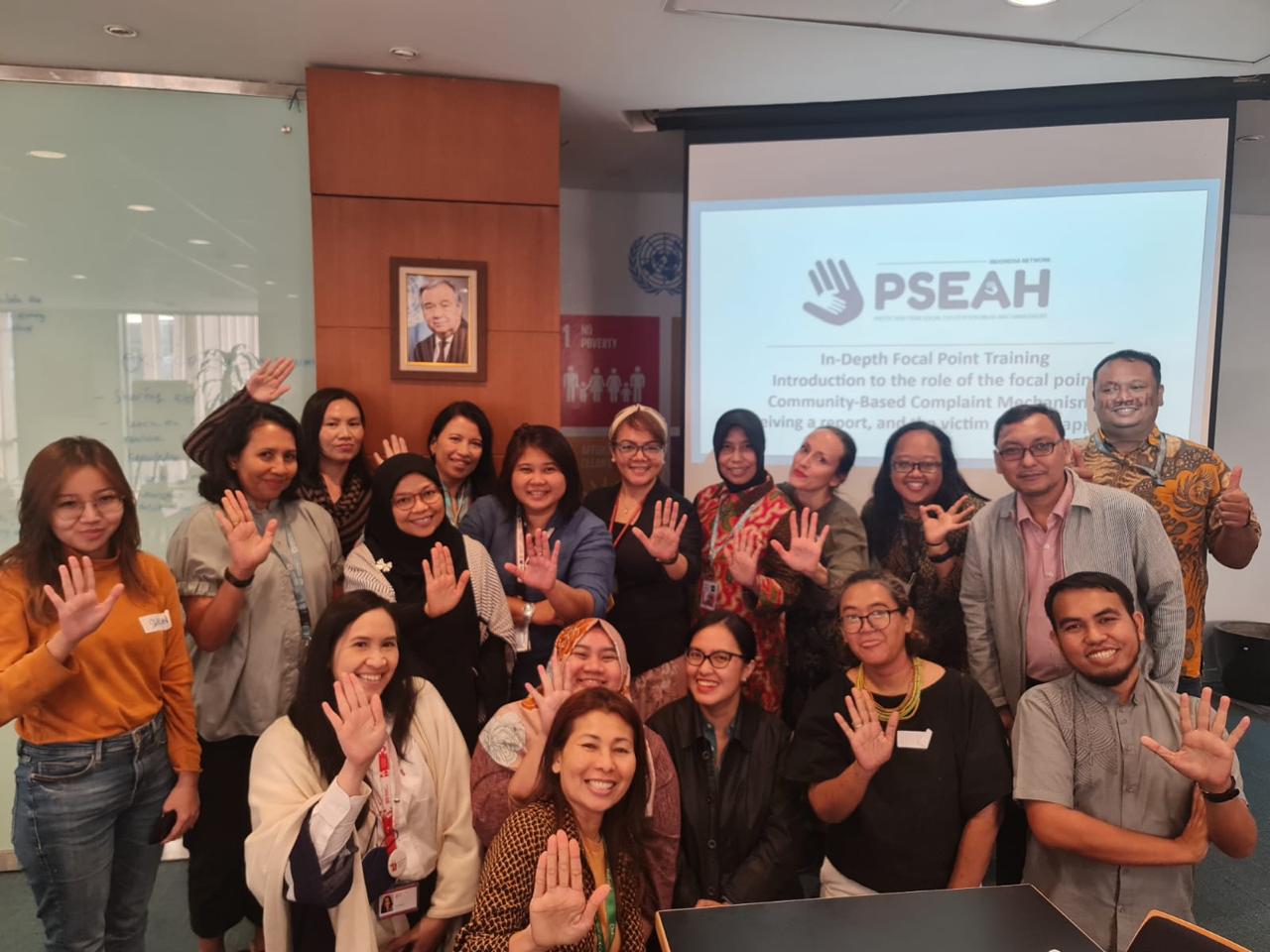 A group of 18 people crouch in front of a screen with their palms displayed. The screen carries mention of prevention of sexual misconduct.