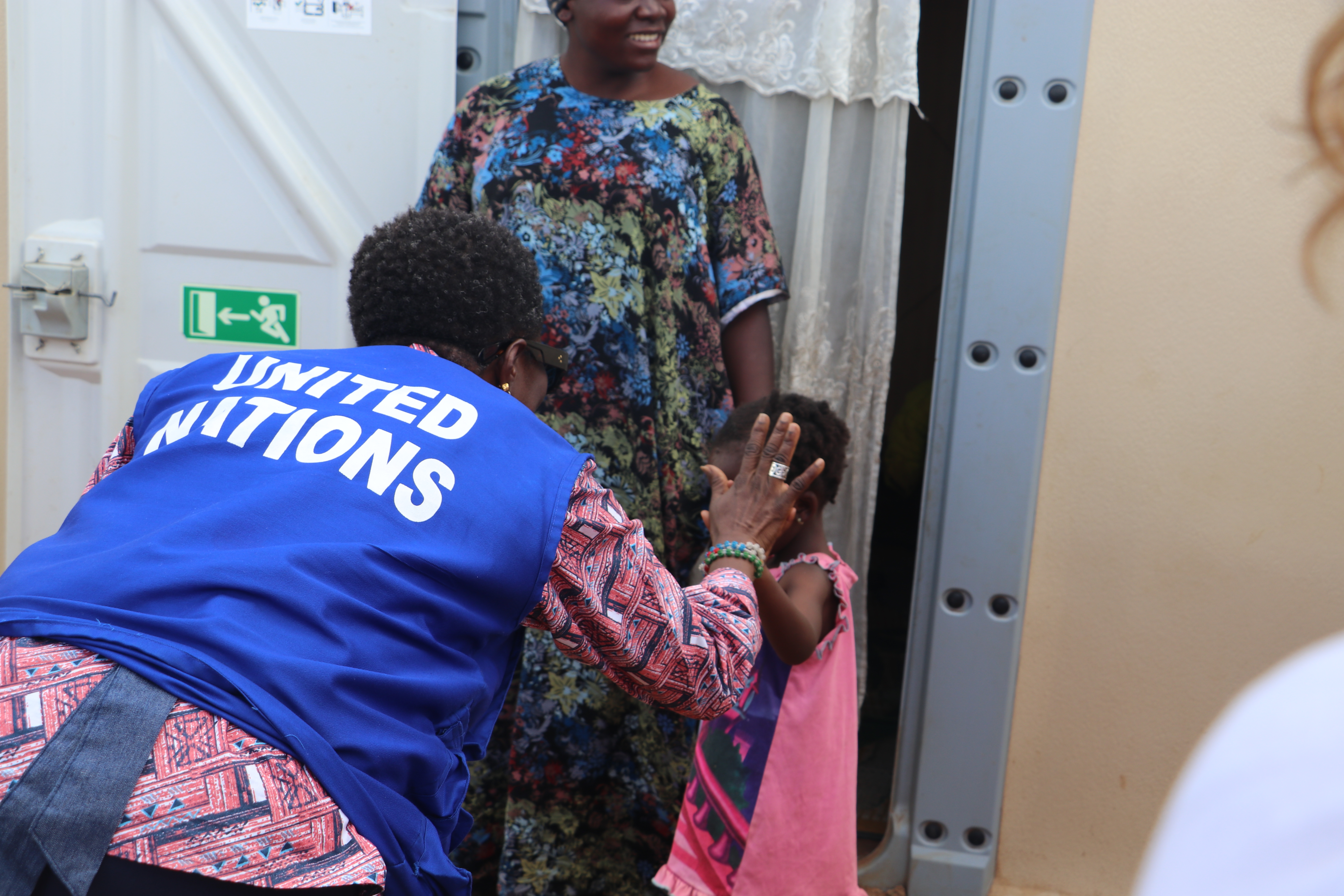 A woman in a red dress and blue vest that says United Nations gives a high-five to a child