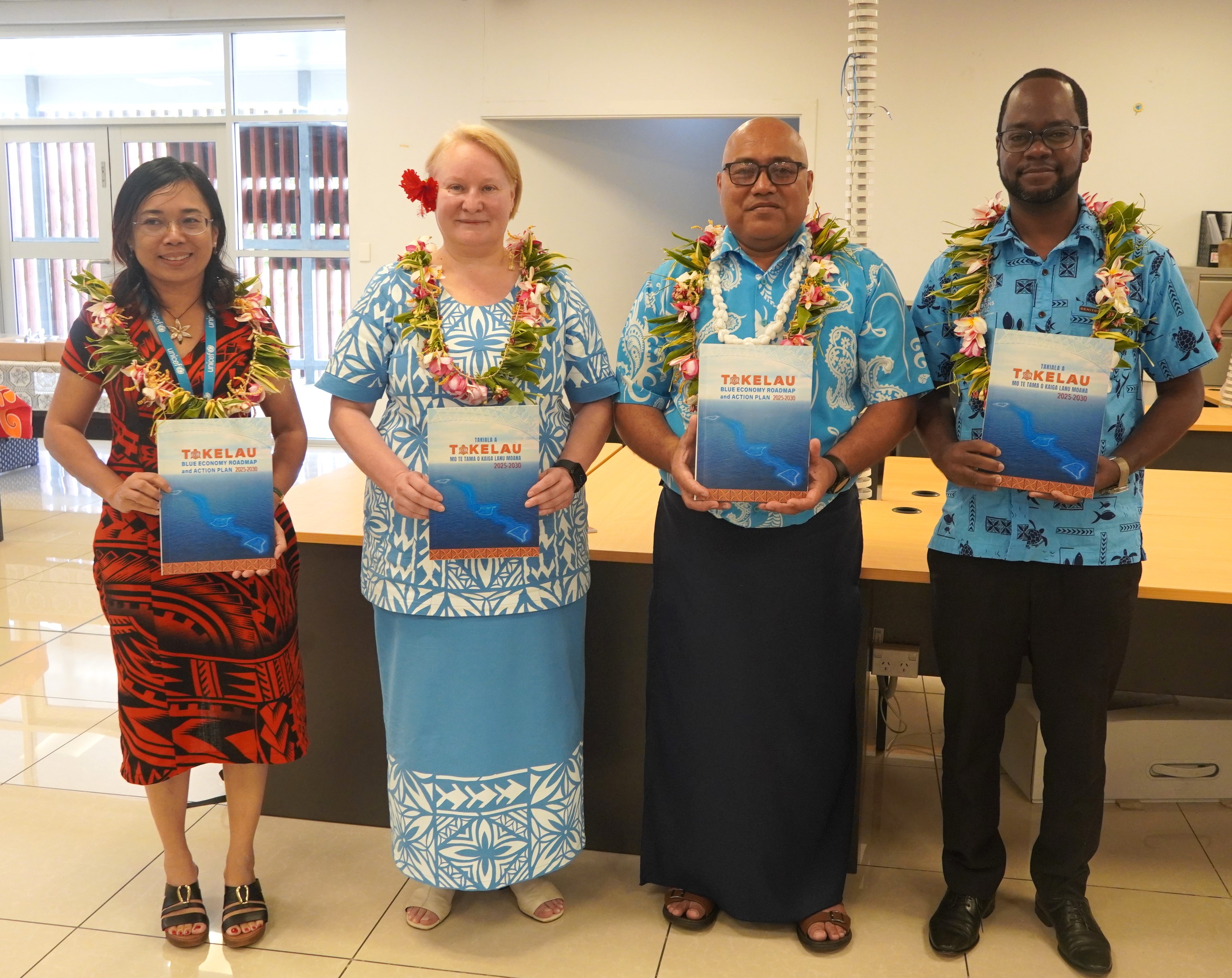 Four people wearing colourful clothing and flower garlands stand in a row, holding the Blue Economy Roadmap