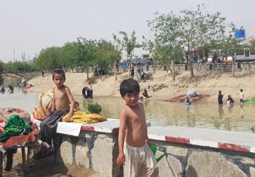 Children stand near a river with people washing clothes and carpets in a water canal.
