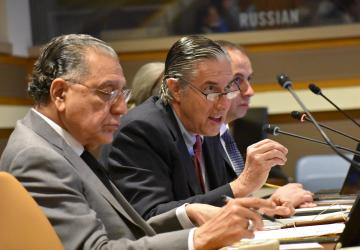 two men in grey suits speak into micro phones at a meeting