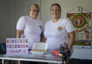 two women in white t-shirts stand behind a kitchen counter 
