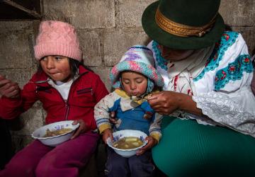 two young children eat a bowel of soup next to their mother 