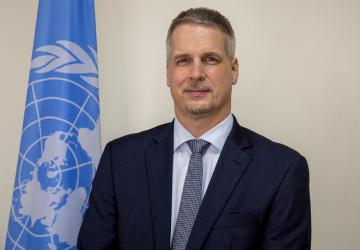 man in dark suit and blue ties stands in front of UN flag 