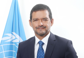 man in dark suit and blue ties stands in front of UN flag 