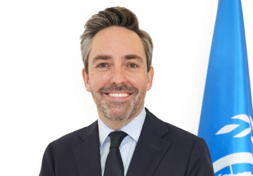 man in dark suit stands next to UN flag against white background 