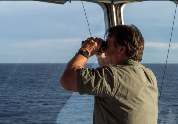 A man in a grey shirt aboard a ship looks out into the sea with binoculars