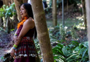 woman with dark hard and traditional indigenous dress stands side ways against a tree 