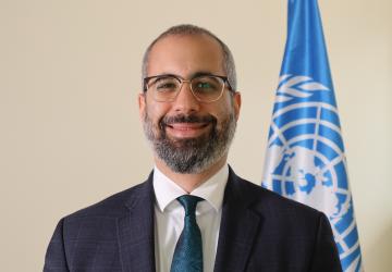 man in dark suit and blue ties stands in front of UN flag 