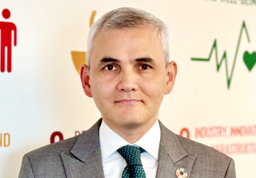 Man in grey suit and green tie stands in front of white wall with SDG symbols 