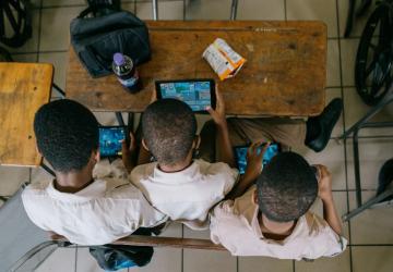view from above of three school children sitting at a desk looking at a tablet 