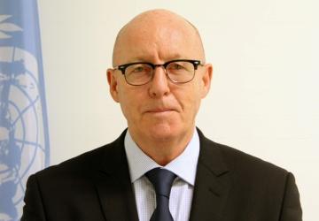 Man in dark suit stands in front of UN flag against white wall looking into the camera