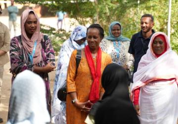 Women in tunics and free-flowing scarves stand in a semi-circle and talk, inside a larger circle of men and women.