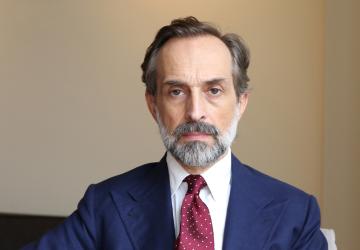 A man in a dark blue suit and red tie and pocket square sitting against a beige background