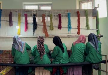 A group of Afghan women sitting on a loom, weaving a colorful rug. They are wearing traditional clothing and head coverings.