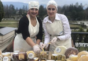 Two individuals in aprons and chef hats arranging a variety of artisan cheeses on a table outdoors