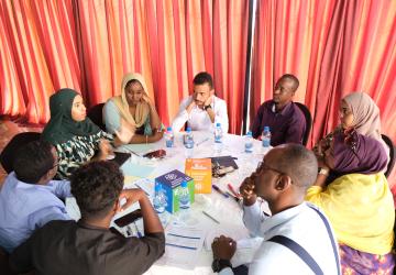 A group of four women and five men gather around a table, in discussion. Papers are on the table along with blocks for the SDG Goals.
