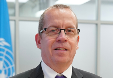 A man wearing glasses stands in front of a grey background with the UN flag behind him. He is wearing a suite, white shirt and tie