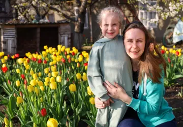 A woman in a green shirt hugs a child in a grey dress with blonde hair in front of a field of flowers