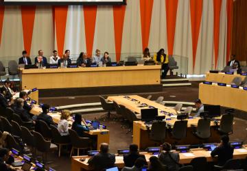 A professional conference room setting with individuals seated at a long table on a stage, facing an audience. Each person at the table has a microphone and papers in front of them, against a backdrop of orange and white vertical stripes. The audience, seated in rows with attached desks and laptops, is attentively engaged in the presentation or discussion taking place.