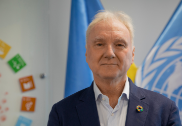 The RC is wearing a black jacket and a blue shirt. He stands in front of the SDG logos and the UN flag