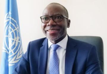 A man is wearing a blue suite, white shirt and blue tie, he is also wearing glasses. There is a UN flag behind him