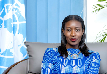 A woman in a blue patterned dress is seated in front of a blue and white background. The UN flag is to her right and a plant is to her left