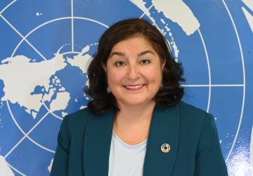 A woman in a bluish green suit and white shirt stands in front of a blue map of the world, likely part of the UN flag