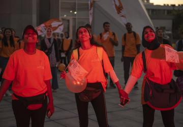 Three young girls are holding hands and looking up at the sky, they are wearing orange the world shirts marking the 16 days of activism against violence against women
