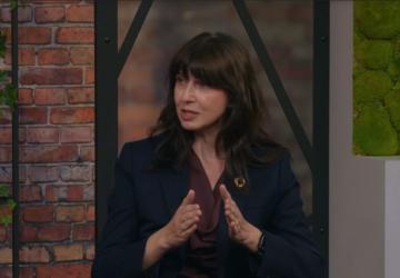 A woman is seated in front of a brickwall background and speaking. She is wearing a black blazer and red top