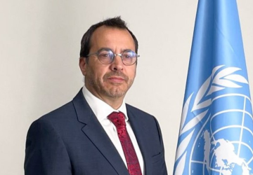 A man wearing a black suite, red tie and white shirt is standing in front of a white background with a 