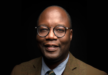 A man stands in front of a black background. He is wearing a brown suite and blue shirt. 