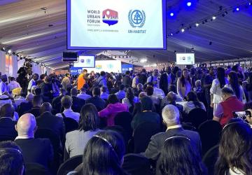 A large group of people in a conference room looking at a board that reads World Urban Forum 