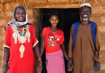 Three people in traditional Eritrean dress look at the camera and smile outside a home