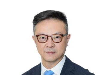 The RC is looking at the camera and behind him is a white background. He is wearing glasses, a dark blue tweed suit, white dress shirt, and light blue tie.