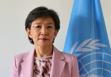 The RC is wearing a pink jacket and standing in front of a grey background with the UN flag