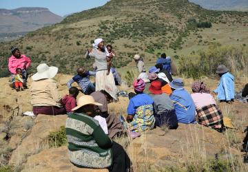 A woman stands in the middle of a group outdoors and speaks to them, while holding her child