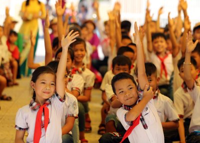 A group of schoolchildren in uniform sit together indoors with many raising their hands.