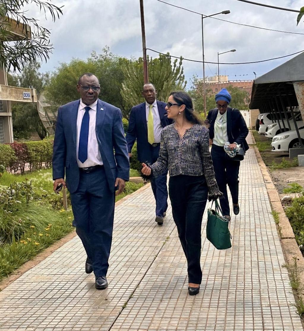 Two people are walking in an outdoor space, one is a man and one is a woman, there are others in the background