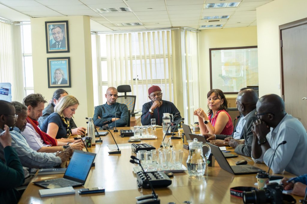 Group of professionals seated around a conference table in a bright meeting room, using laptops and notebooks while participating in a discussion; microphones, water glasses, and documents are arranged on the table, with framed posters and a map visible on the walls behind them.