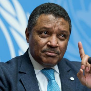 A man in a blue tie points upward while speaking into a microphone.