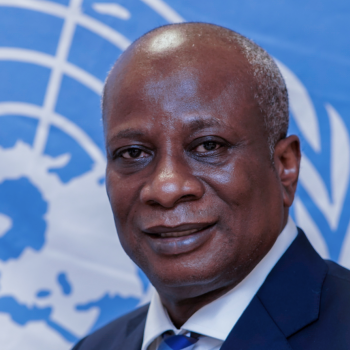 A man with a kind face smiles at the camera in front of the United Nations logo. 