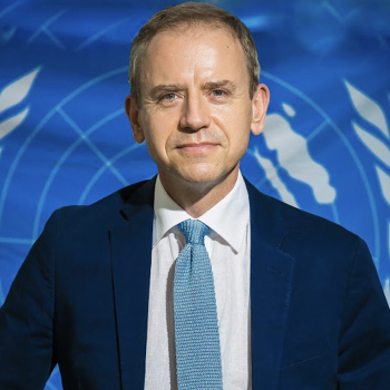 A man with a light blue tie and dark blue suit jacket looks at the camera.