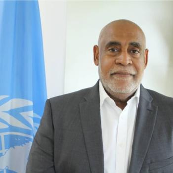 A man stands next to the United Nations Flag. 