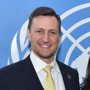 A man in a yellow tie and white shirt smiles at the camera in front of the United Nations logo.