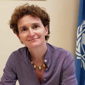 A woman in a purple shirt smiles at the camera near a United Nations flag. 