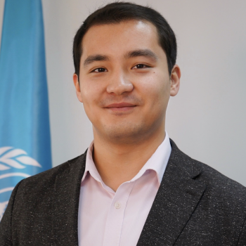 A man in a white shirt and black suit stands next to the United Nations flag.