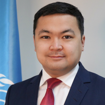 An elegant man in a red tie and white shirt stands next to the United Nations flag.