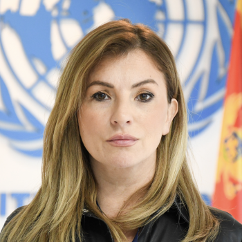 A woman with long hair looks straight at the camera in front of the United Nations logo. 