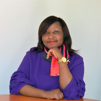 women in purple suit and pink scarf sits down at a table 